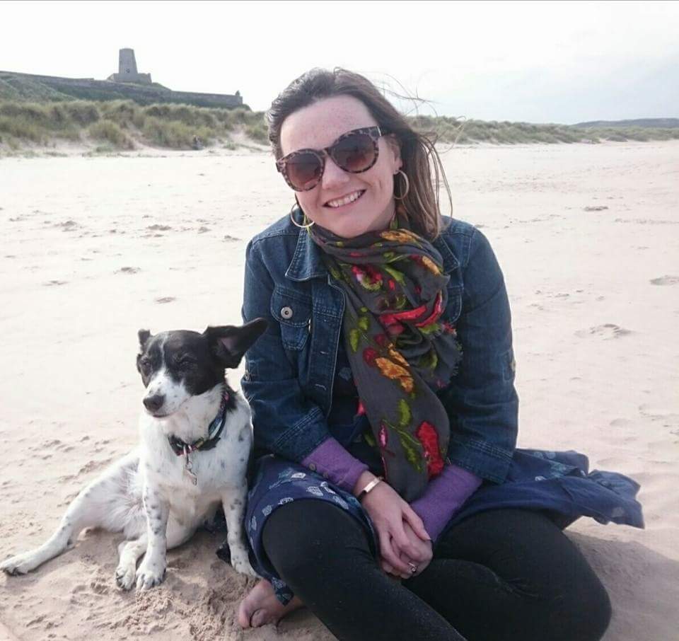 Lisette, a white woman with freckles and brown hair, is sitting barefoot on a beach. She is smiling widely and is wearing sunglasses, a scarf, denim jacket and blue dress with leggings. Harper Lee, her black and white little rescue dog sits and leans beside her, her ears are sticking up because it's windy. Bamburgh Castle sits behind them on the horizon.