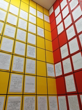 photo description: really high red and yellow walls with black grid outlines filled with white posters of words