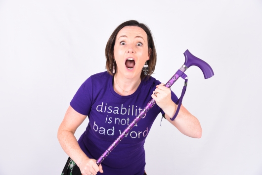 Image shows Lisette in a purple t-shirt which reads disability is not a bad word, holding her walking stick with her eyes and mouth wide open.